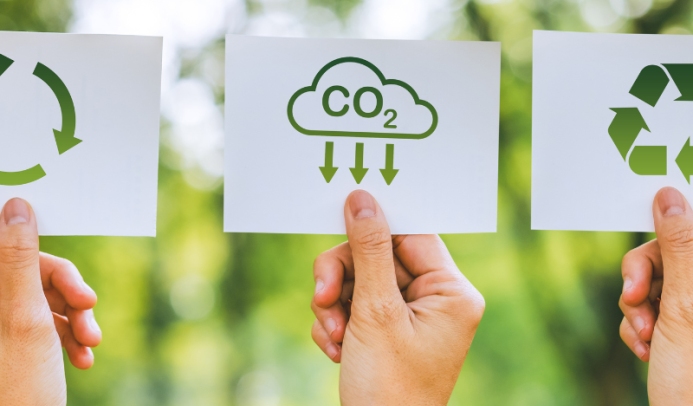 Three hands holding cards against a blurred green background. The cards display green environmental icons: on the left, a circular recycling symbol; in the middle, a cloud labeled “CO₂” with arrows pointing downward, representing carbon capture or reduction; and on the right, a recycling triangle symbol. This image conveys themes of sustainability and environmental responsibility.