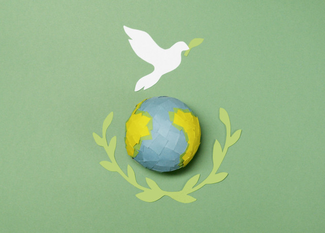 A stylized image of a globe crafted with blue and yellow paper, surrounded by olive branches on either side, symbolizing peace. Above the globe, a white dove holds an olive branch in its beak, set against a green background. This composition conveys themes of peace, unity, and environmental harmony.