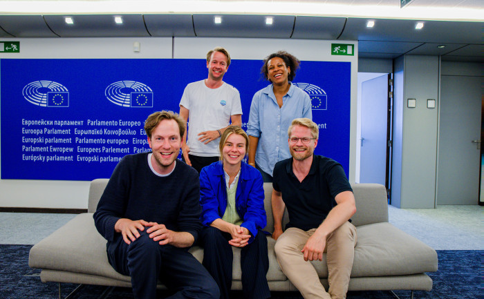 Group photo of five MEPs from Volt Europa at the European Parliament. They are seated on a light-colored couch in front of a blue wall with the European Parliament logo and multilingual text that reads “European Parliament.” From left to right: seated in front are Damian, Anna, and Kai, while Reinier and Nela stand in the back, all smiling. The setting is modern and well-lit.