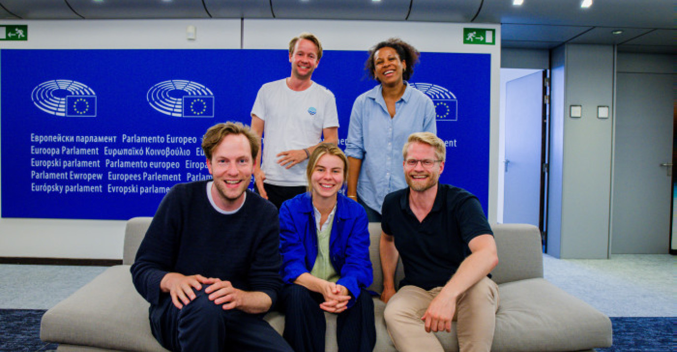 Group photo of five MEPs from Volt Europa at the European Parliament. They are seated on a light-colored couch in front of a blue wall with the European Parliament logo and multilingual text that reads “European Parliament.” From left to right: seated in front are Damian, Anna, and Kai, while Reinier and Nela stand in the back, all smiling. The setting is modern and well-lit.