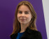 Portrait of Anna, with a gentle smile, wearing a black jacket over a blue top. She has long, light brown hair and stands in front of a purple and white background.