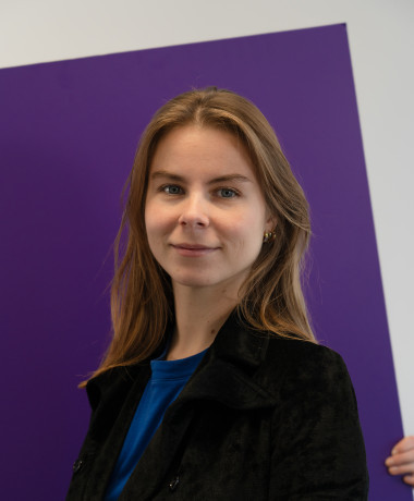 Portrait of Anna, with a gentle smile, wearing a black jacket over a blue top. She has long, light brown hair and stands in front of a purple and white background.