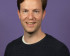 Portrait of Damian, smiling and wearing a dark sweater over a white shirt. He has short, wavy hair and stands against a solid purple background.