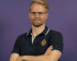 ortrait of Kai, standing with arms crossed and wearing a dark polo shirt with a small embroidered emblem. He has short blonde hair, glasses, and a slight smile, set against a solid purple background.