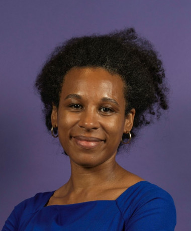 Portrait of Nela, smiling and wearing a royal blue top. She stands against a solid purple background, with her hair styled naturally and wearing small hoop earrings.