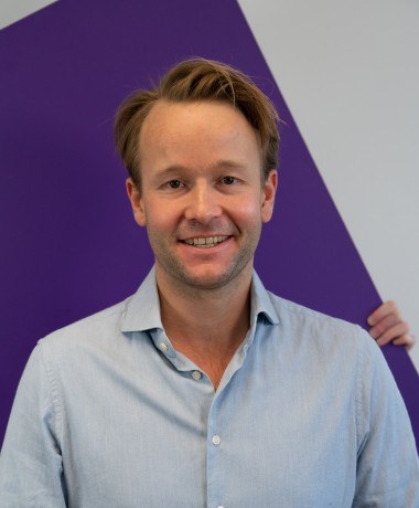 Portrait of Reinier van Lanschot, smiling and wearing a light blue button-down shirt. He stands against a background featuring a large purple geometric shape.