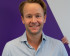 Portrait of Reinier van Lanschot, smiling and wearing a light blue button-down shirt. He stands against a background featuring a large purple geometric shape.